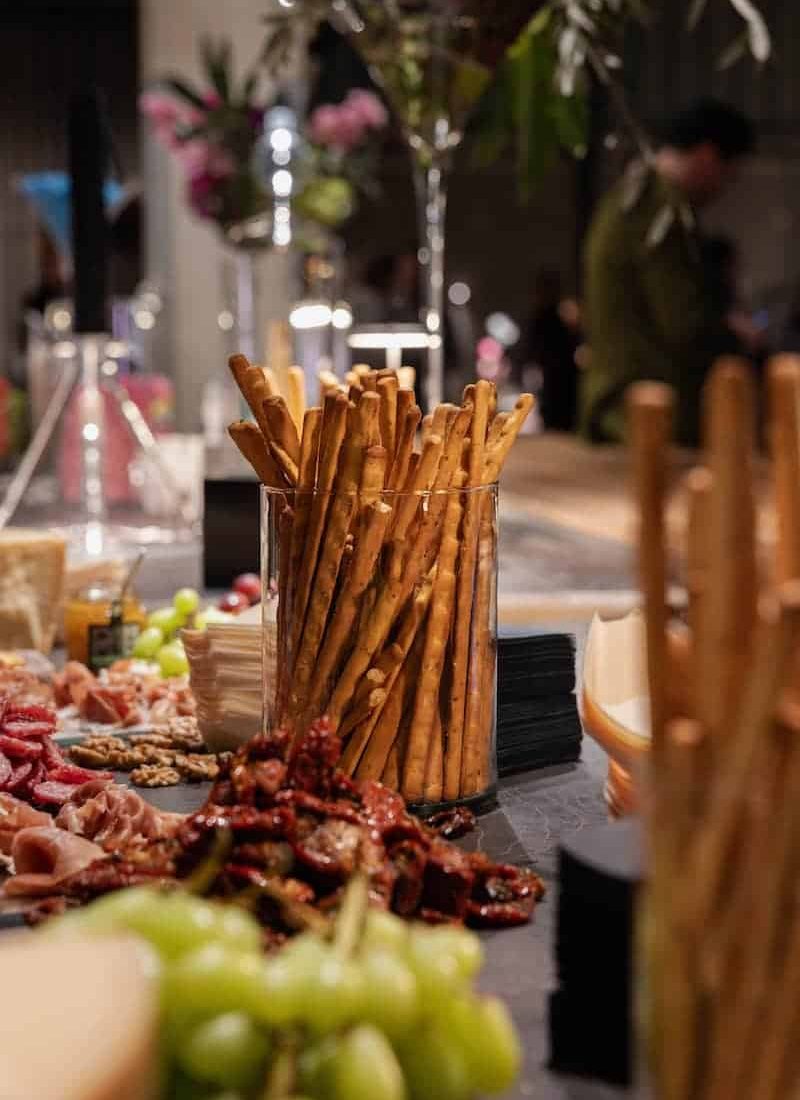 Ein mit Grissini gefüllter Glasbehälter ist von verschiedenen Lebensmitteln umgeben, darunter Weintrauben, Nüsse und Fleisch, die auf einem Tisch angeordnet sind. | casaceramica