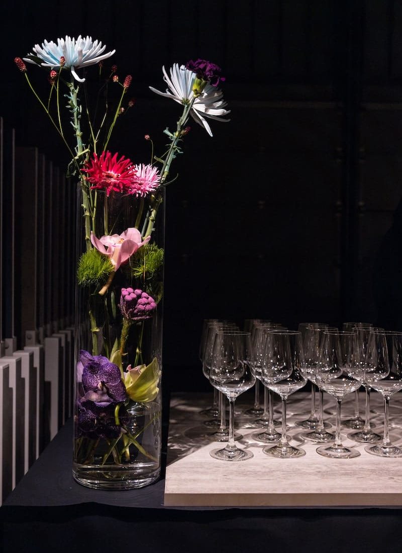 Eine große, durchsichtige Vase mit verschiedenen Blumen steht neben einem Tablett mit leeren Weingläsern auf einem dunklen Tisch. | casaceramica