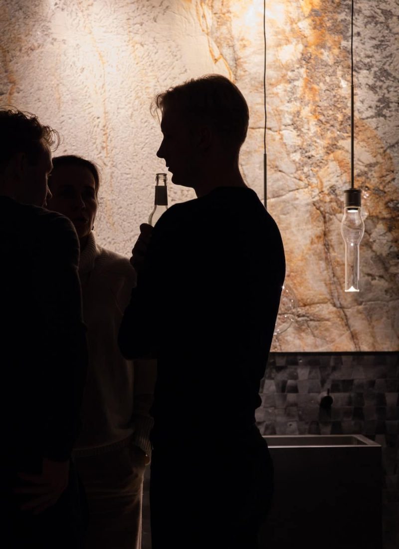 Die Silhouette einer Person, die eine Flasche hält, steht vor einer strukturierten Wand mit hängenden Glühbirnen. Im Hintergrund sind teilweise zwei weitere Personen zu sehen. | casaceramica