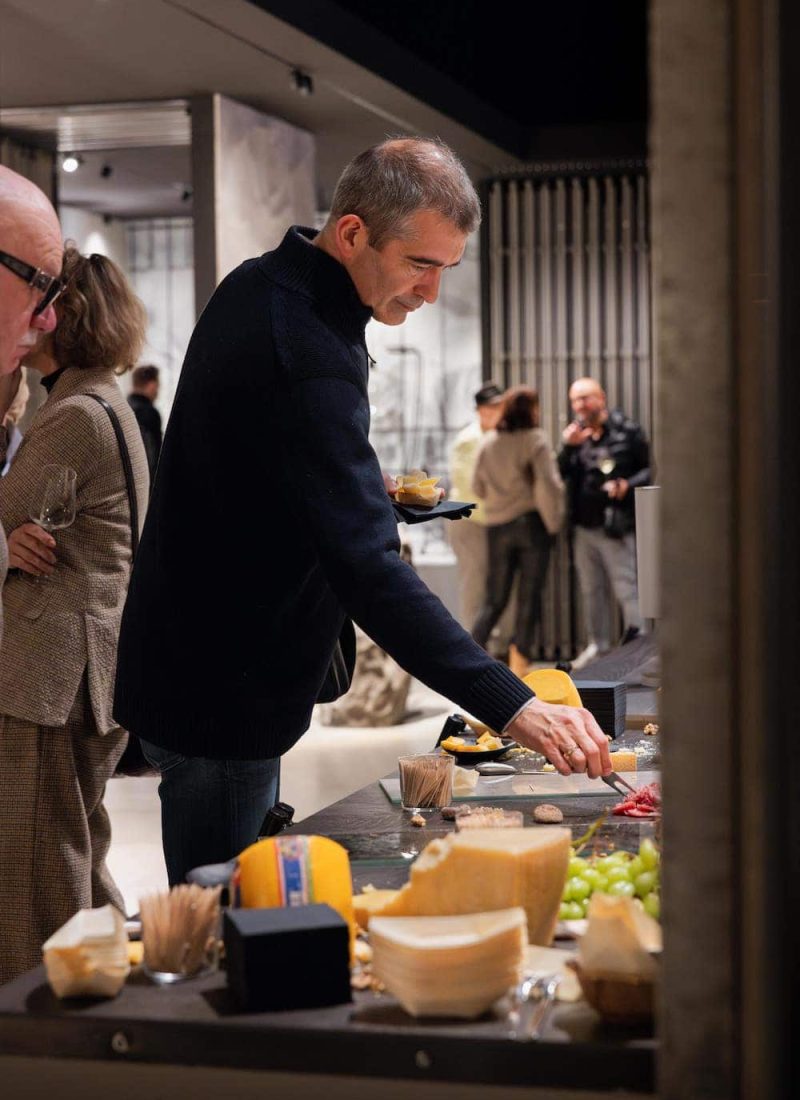Ein Mann in einem dunklen Pullover nimmt sich bei einer Veranstaltung Essen von einem Tisch, auf dem Käse, Trauben und andere Dinge liegen. Im Hintergrund sind weitere Personen zu sehen. | casaceramica