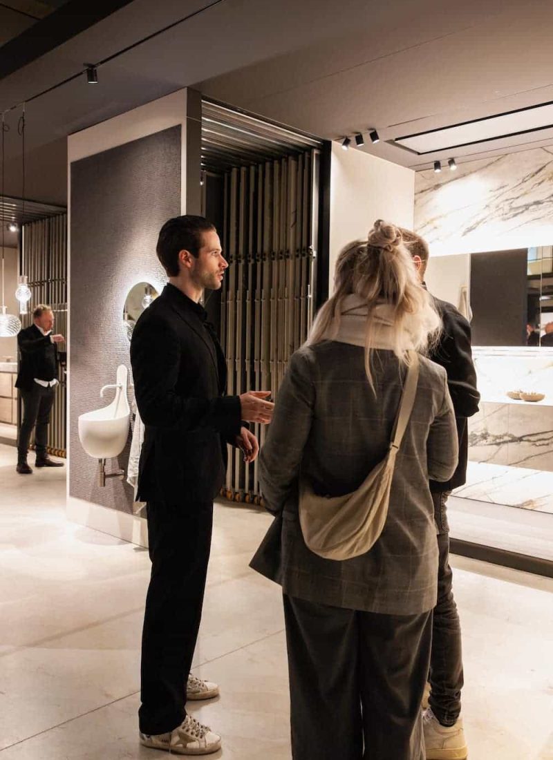 Drei Personen unterhalten sich in einem modernen, gut beleuchteten Ausstellungsraum. Im Hintergrund sind verschiedene Einrichtungsgegenstände zu sehen, darunter eine Marmorwand und ein Waschbecken. | casaceramica