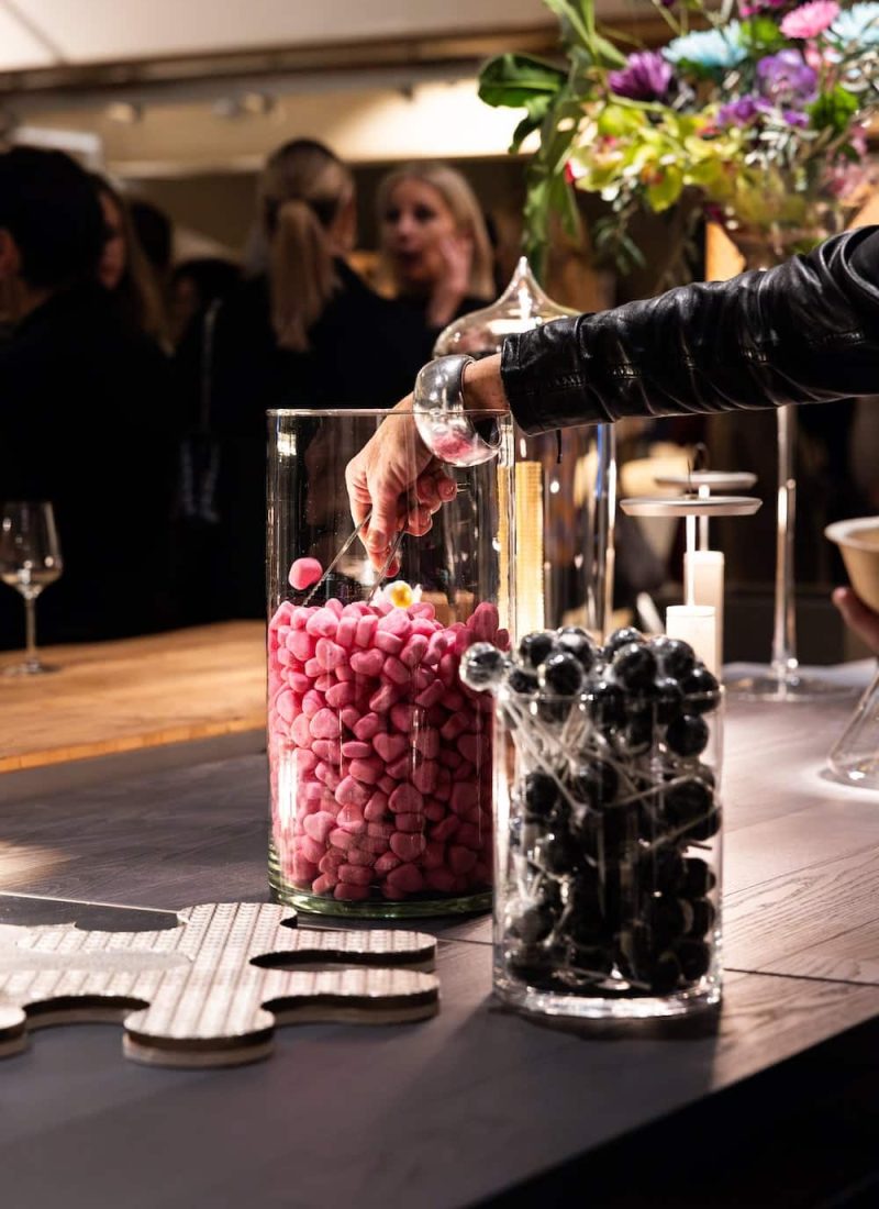 Eine Person greift in ein Glasgefäß mit rosafarbenen Bonbons auf einem Tisch. Ein weiteres Glas mit schwarzen Bonbons und Deko-Elementen ist ebenfalls zu sehen. Im Hintergrund sind ein paar Personen und Blumenarrangements zu sehen. | casaceramica