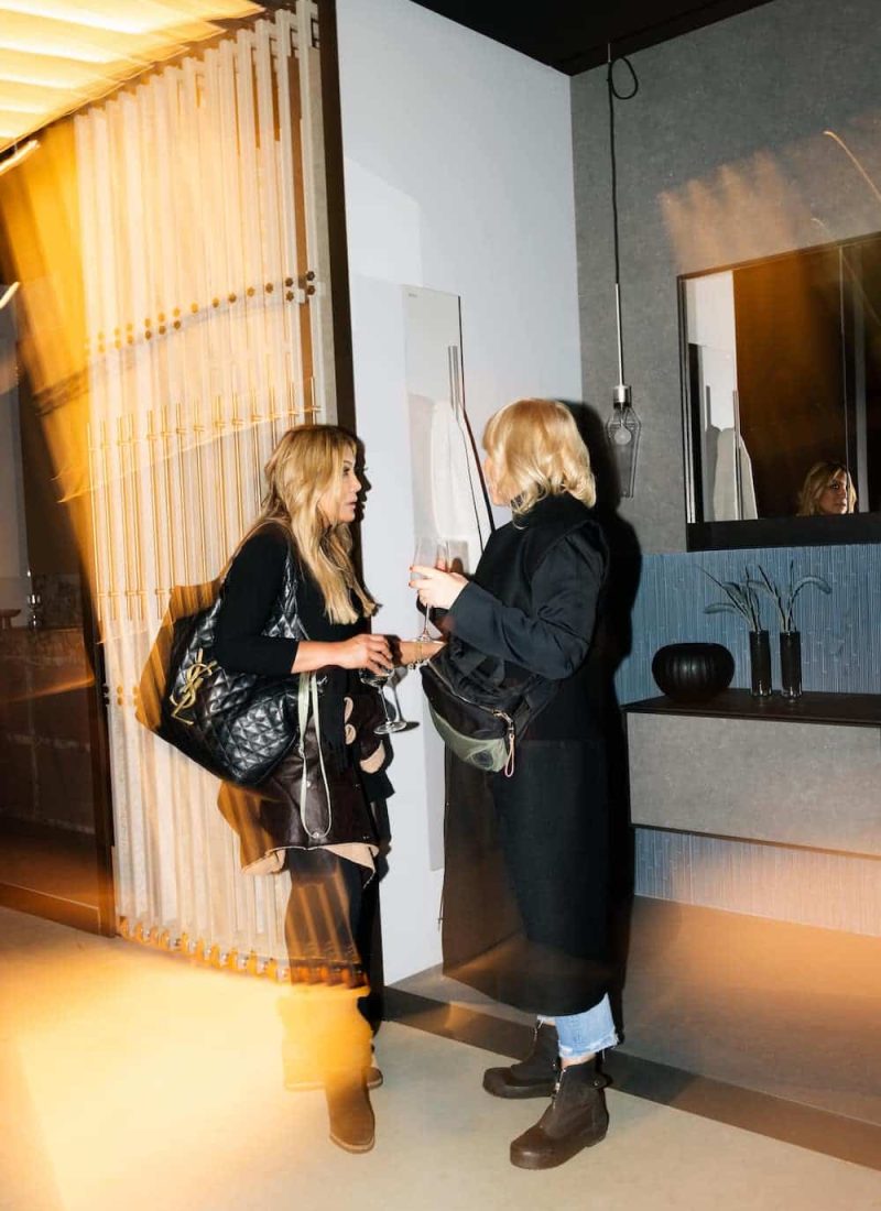 Zwei Personen stehen im Gespräch und halten Getränke in der Hand. Beide sind dunkel gekleidet. Im Hintergrund ist ein modernes, minimalistisches Interieur mit warmer Beleuchtung zu sehen. | casaceramica