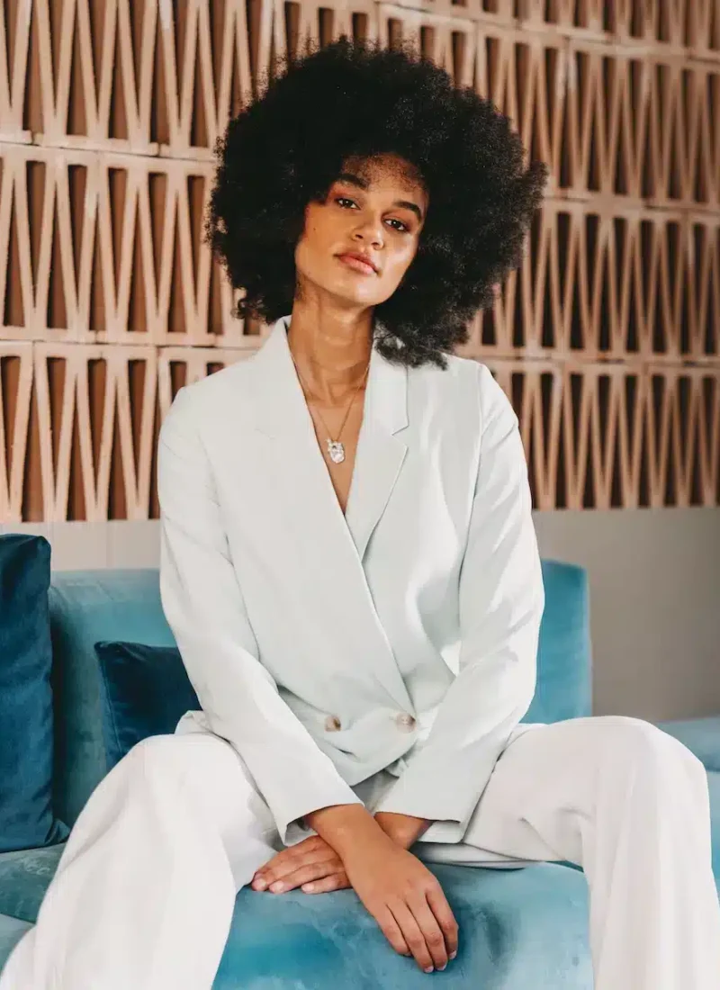 Frau mit Afrofrisur sitzt auf einem blauen Sofa. Sie trägt einen hellen Blazer und eine Hose und blickt in entspannter Pose in die Kamera. Im Hintergrund ist eine gemusterte Wand mit dreieckigen Ausschnitten zu sehen. | casaceramica