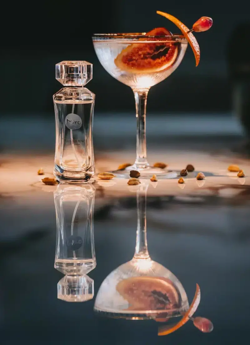 Convary Neben einem Cocktail steht ein Parfümflakon, garniert mit einer Zitrusscheibe und einer Kirsche. Beide spiegeln sich auf einer glänzenden Oberfläche. Auf der Oberfläche sind verstreute Samen zu sehen. | casaceramica