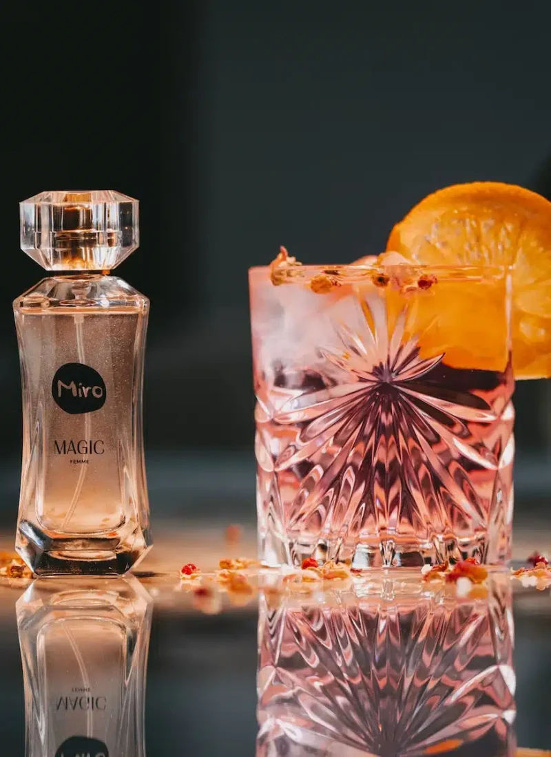Eine Glasflasche mit Miro Magic-Parfüm steht neben einem Kristallglas, das mit einem rosa Getränk und Eis gefüllt und mit einer Orangenscheibe garniert ist. Glas und Flasche stehen auf einer reflektierenden Oberfläche. | casaceramica