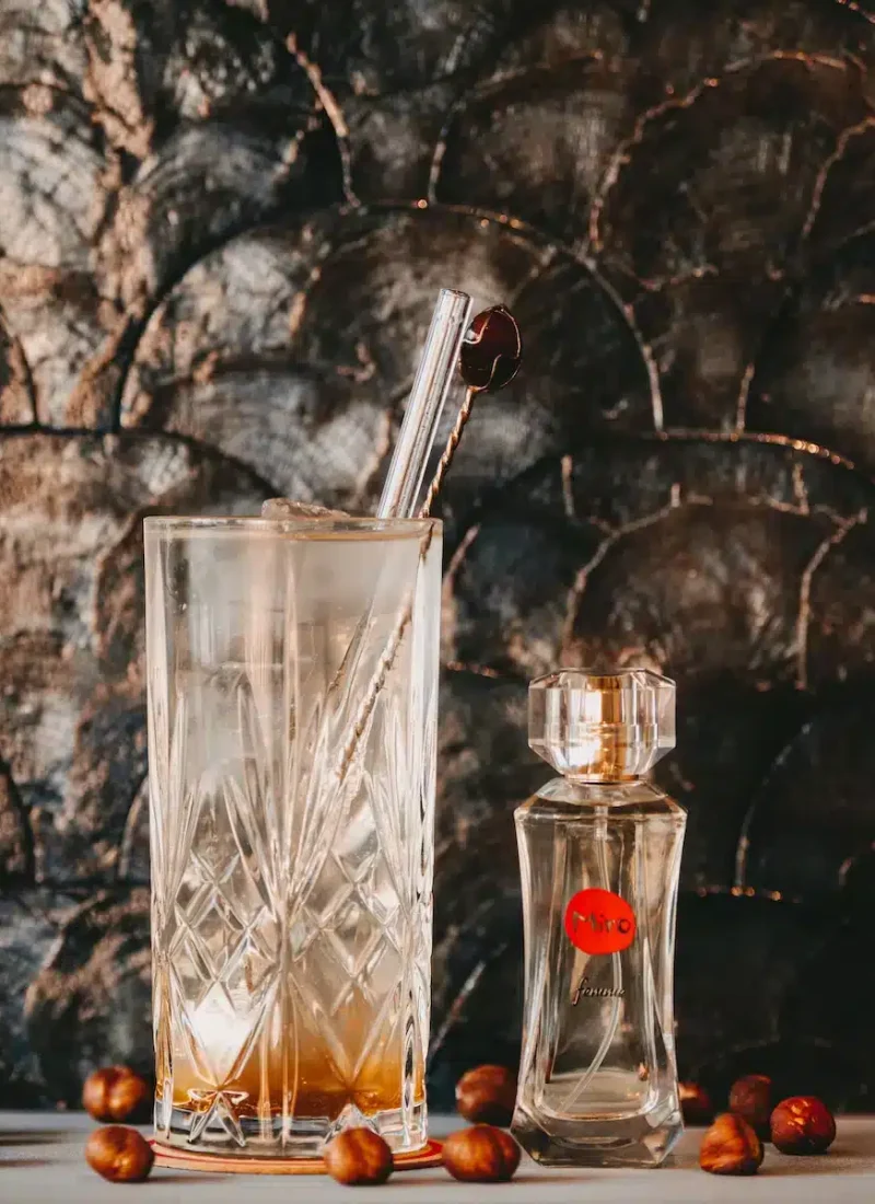 Ein Kristallglas mit einem eisgekühlten Getränk und einem Glasstrohhalm neben einer Parfümflasche auf einem Tisch, mit einem dunklen strukturierten Hintergrund und verstreuten Haselnüssen um die Präsentation herum. | casaceramica