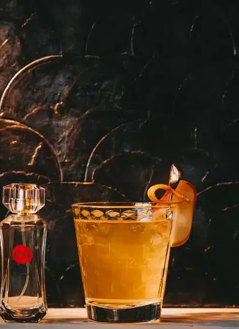 Convary Ein mit einer Orangenschale garniertes Whiskyglas steht neben einer kleinen Glasflasche auf einem Tisch. Der Hintergrund ist dunkel mit leuchtenden Mustern. | casaceramica