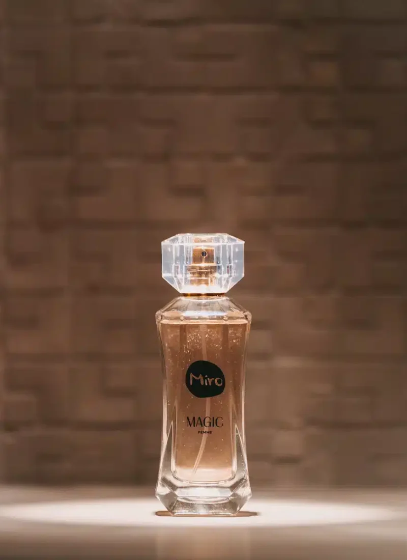 Convary Vor einem strukturierten Hintergrund ist eine durchsichtige Flasche Miro Magic-Parfüm zu sehen. Die Flasche hat einen achteckigen Verschluss und eine goldfarbene Flüssigkeit im Inneren. | casaceramica