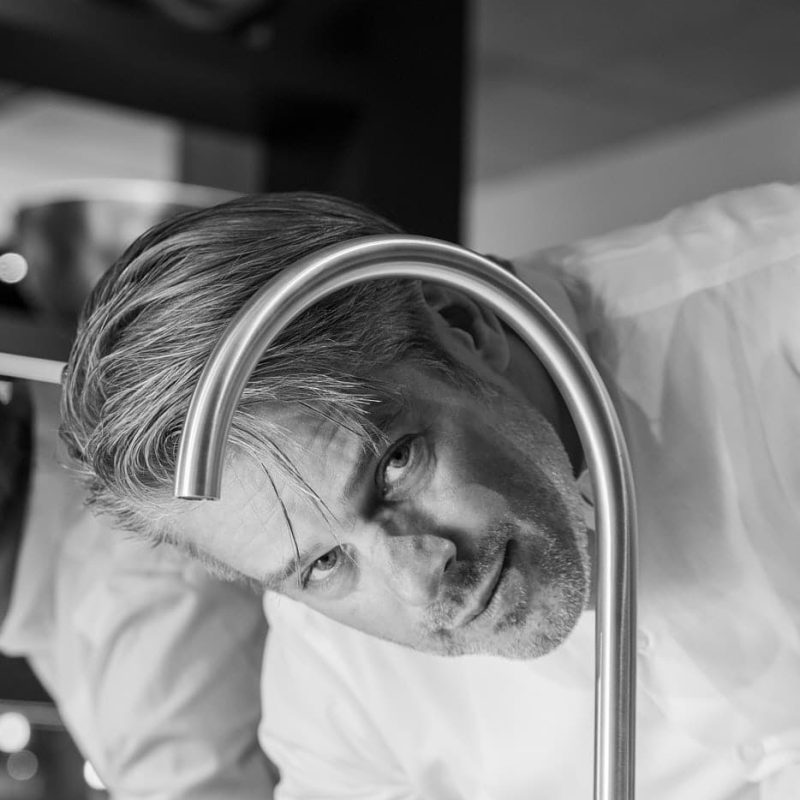 Ein Mann im weißen Hemd blickt aufmerksam unter einen Edelstahlhahn in einer Küche. | casaceramica
